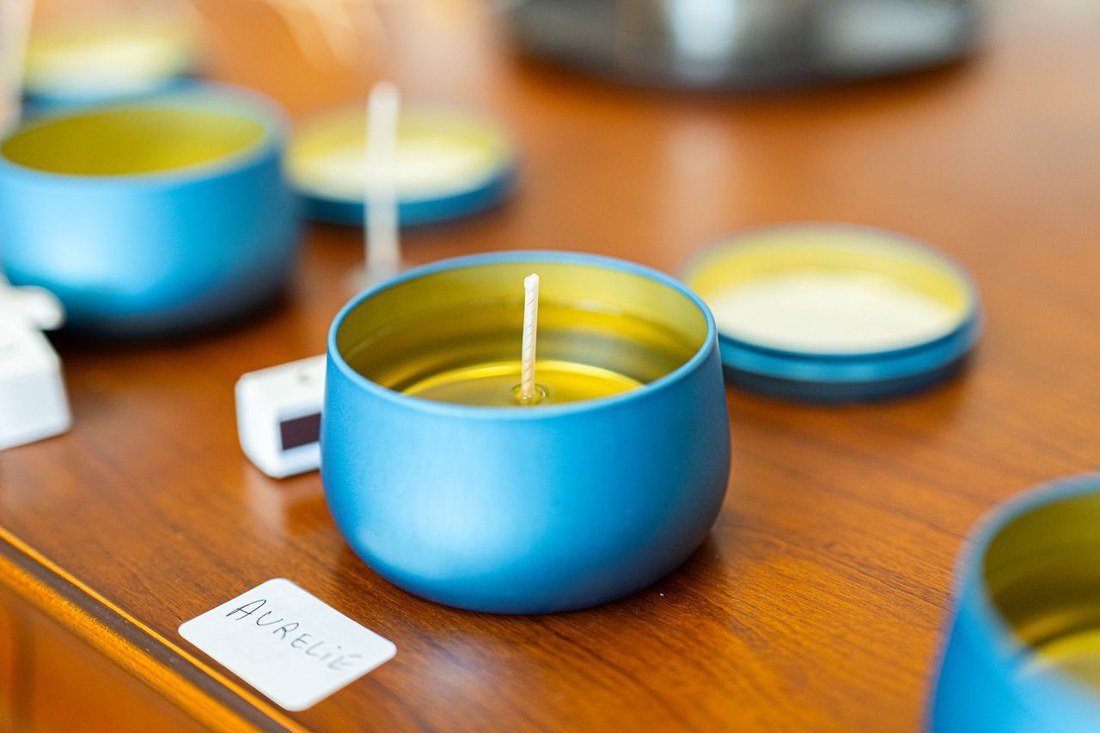 Une boîte métal bleue remplie de cire encore liquide, la mèche de coton bien droite au centre, sur la table de l'atelier — devant elle, l'étiquette manuscrite au prénom de la participante.