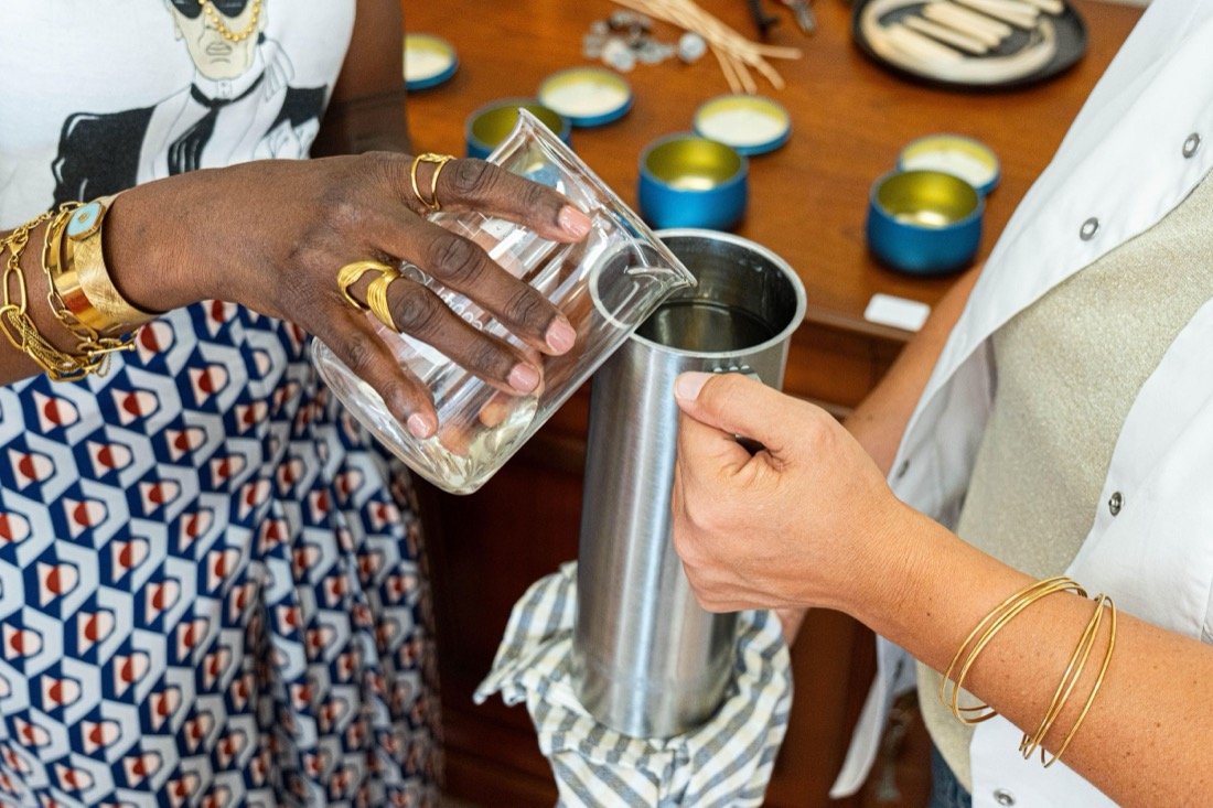 Deux mains versent un parfum transparent d'un bécher de verre dans un pichet métallique, devant les pots de cire déjà coulés.