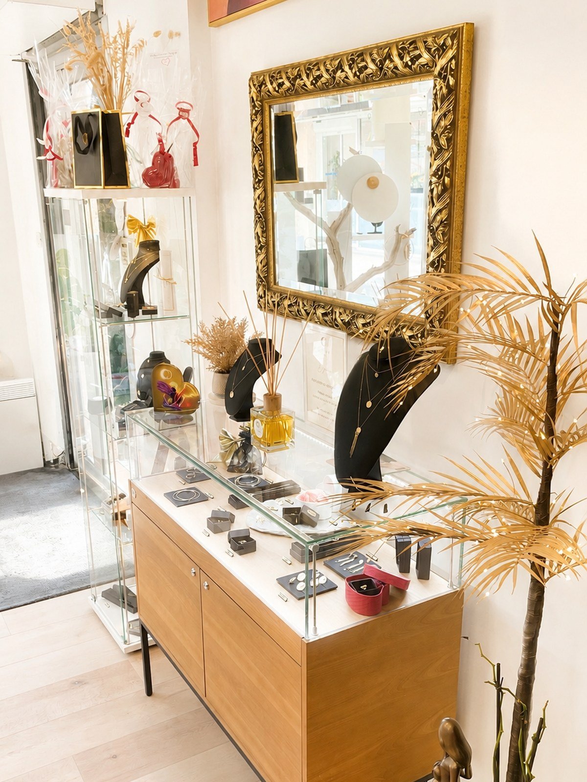 Le comptoir des bijoux sous un miroir dore : bagues, bracelets et colliers presentes sous verre, deux bustes noirs portant des chaines fines, un flacon de parfum et un palmier dore.