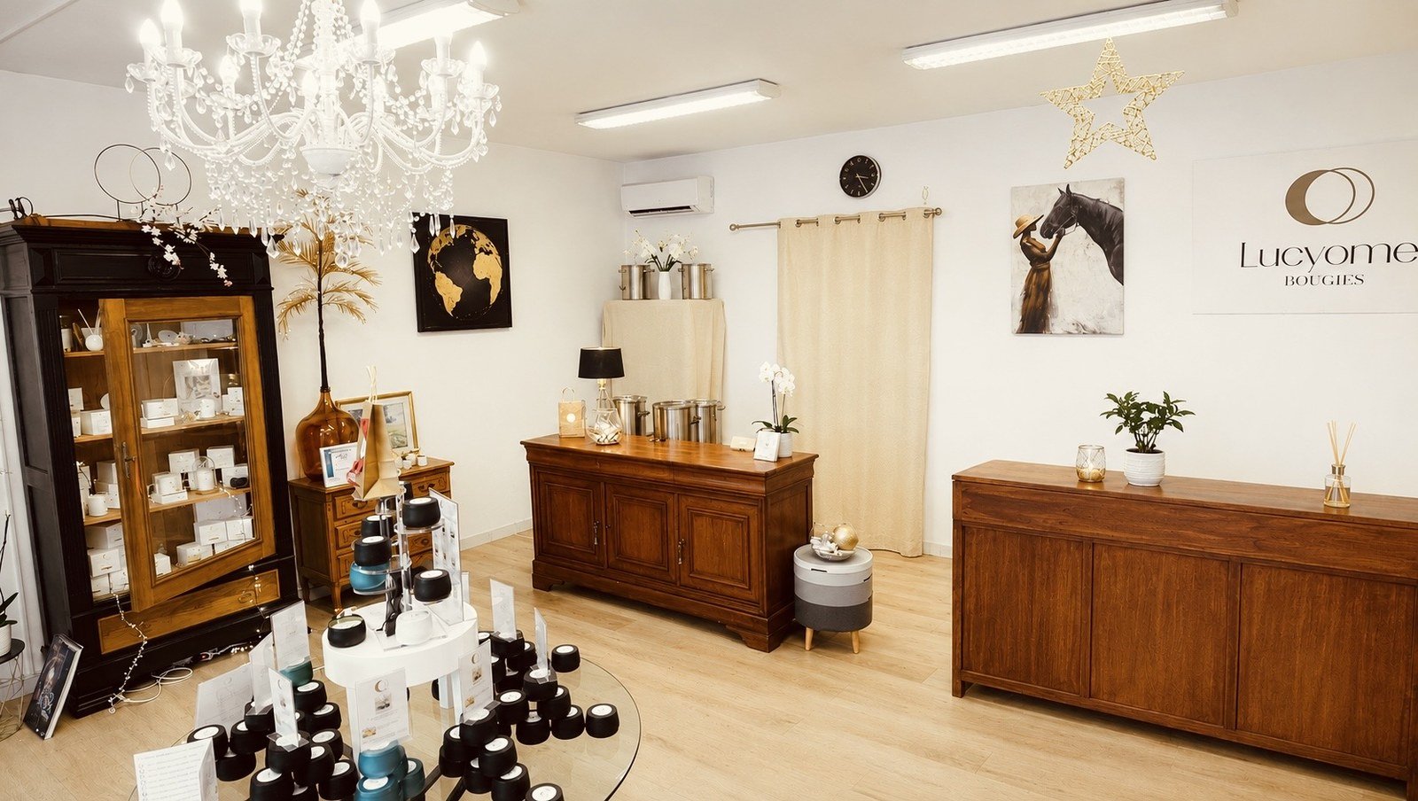 La salle de la boutique Lucyome sous son lustre de cristal : la vitrine noire garnie de bougies et de coffrets, la table de verre ou les Lucyometes sont empilees avec leurs cartes, les buffets de bois le long des murs blancs et le panneau Lucyome Bougies au fond a droite.